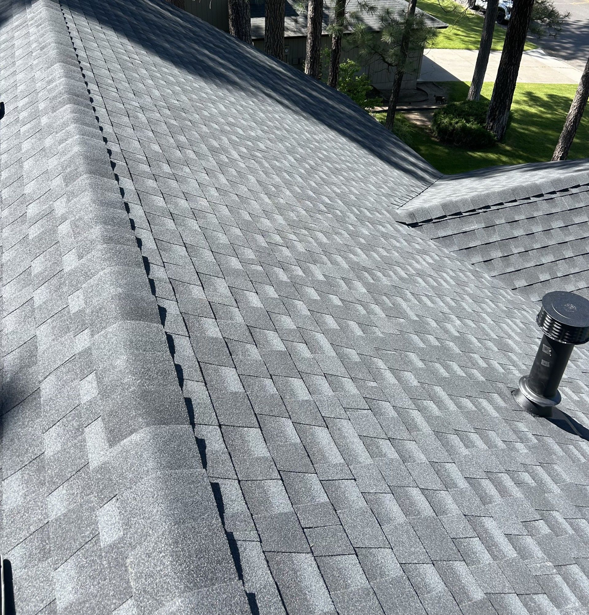A high-angle view of a grey shingled roof with a black plumbing vent pipe and pine trees in the background.