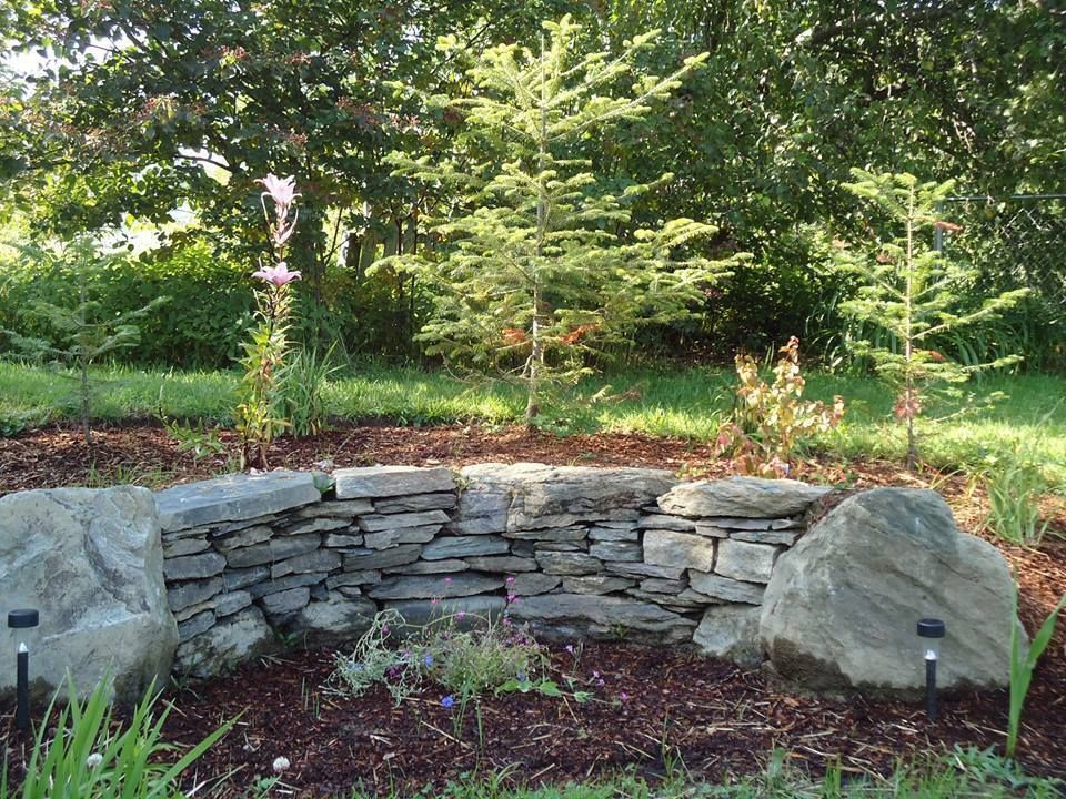 Stone wall with garden bed, surrounded by mulch, two boulders, and greenery.