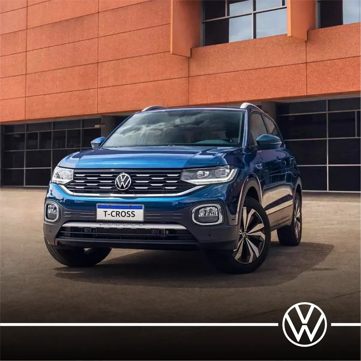 SUV Volkswagen T-Cross azul estacionado em frente a um prédio de tijolos. O logotipo da VW está no canto inferior direito.