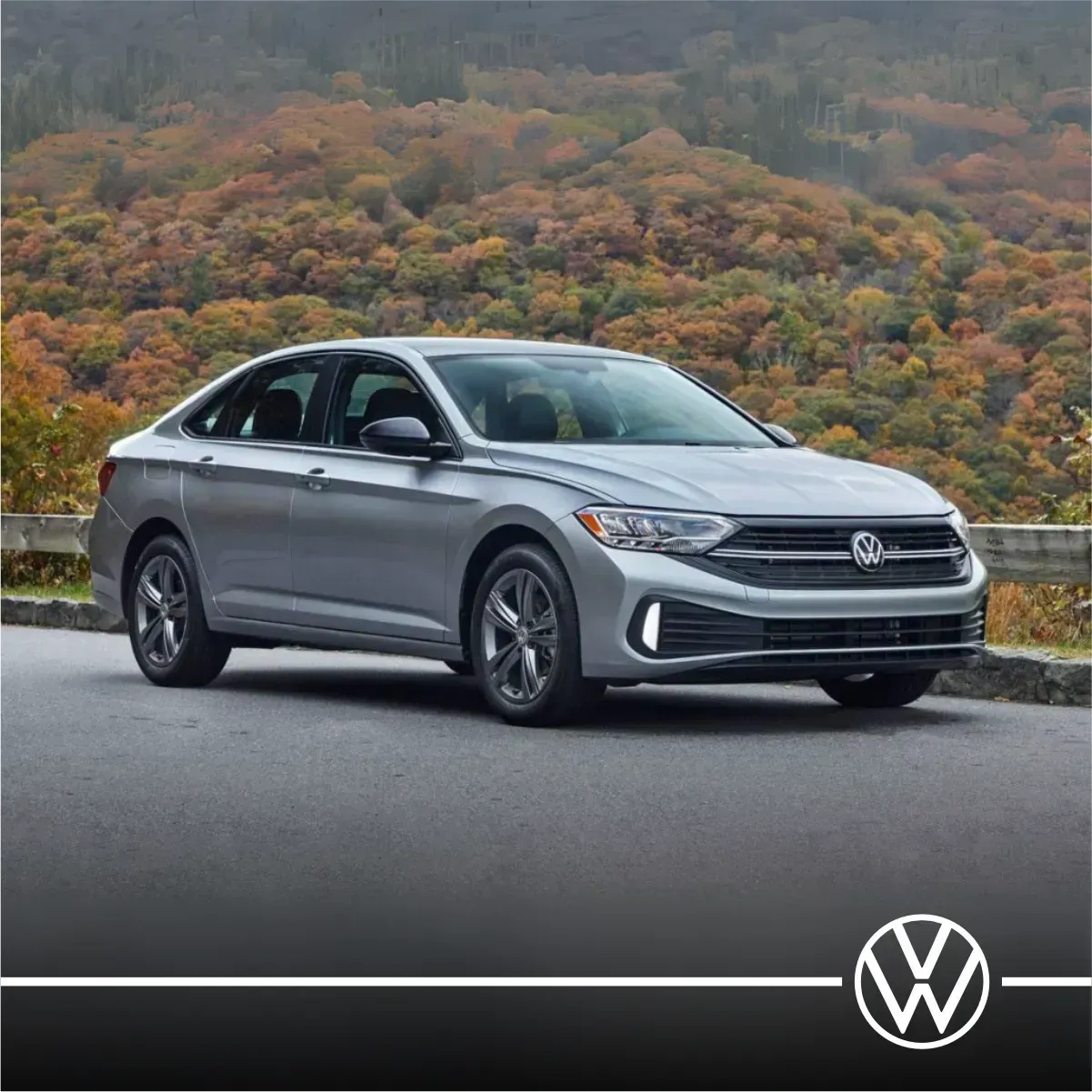 Sedan Volkswagen Jetta prateado em uma estrada com folhagem de outono ao fundo.