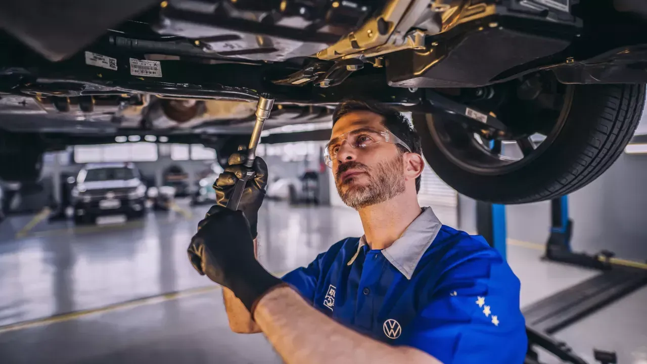 Mecânico trabalhando embaixo de um carro, usando óculos de segurança e luvas em uma oficina mecânica.