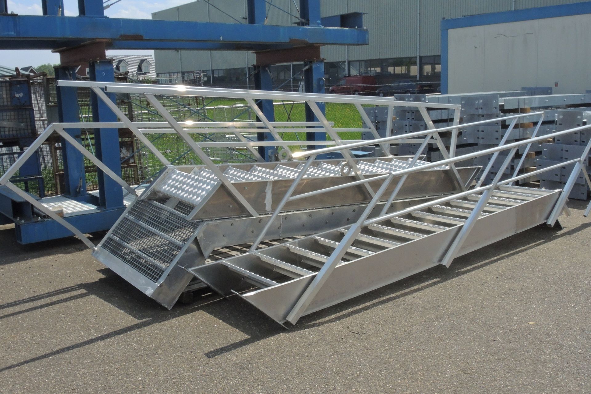 Twee industriële, zilverkleurige metalen trappen met leuningen, die buiten op een grindoppervlak staan.