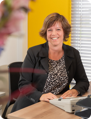 Een vrouw zit aan een bureau met een printer en glimlacht. Colinda Hol