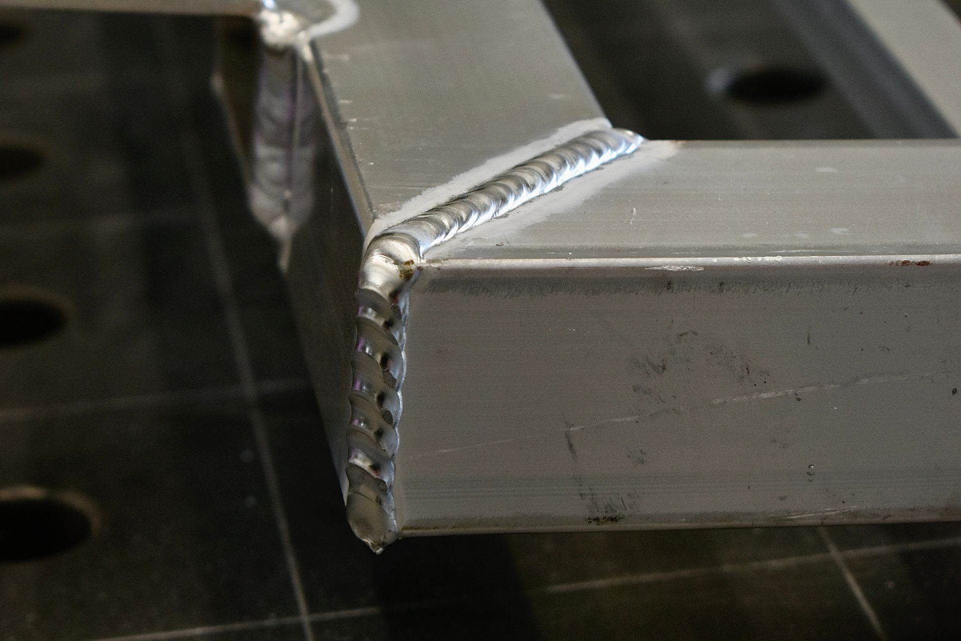 Een close-up van een metalen hoekverbinding met strakke, gelaste naden op een vierkante stalen buis die op een rastertafel rust.