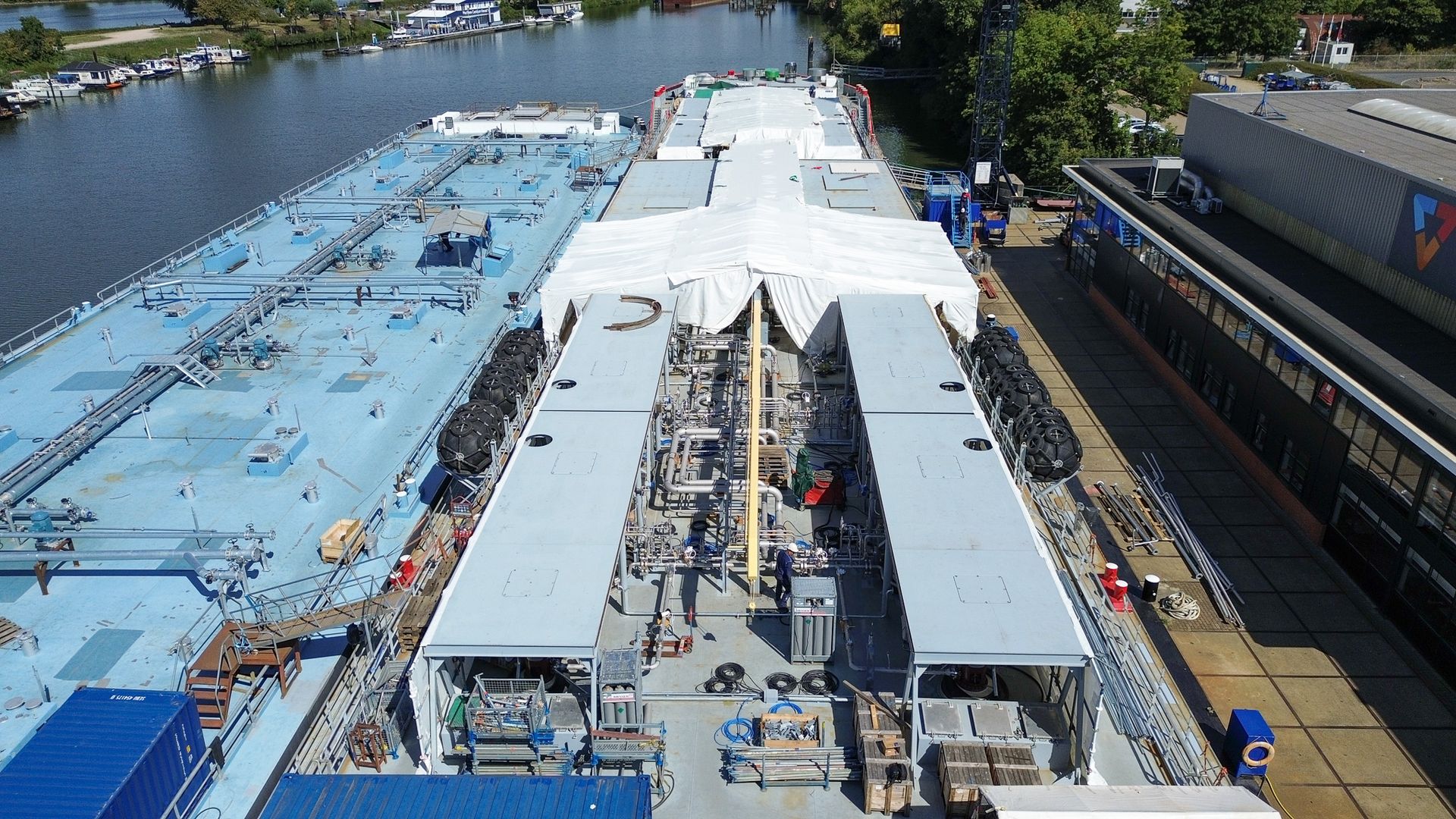 Een opname vanuit een hoog perspectief van een drijvende evenementenstructuur op een rivier, met een blauw dek, witte tenten en industrieel bouwmateriaal
