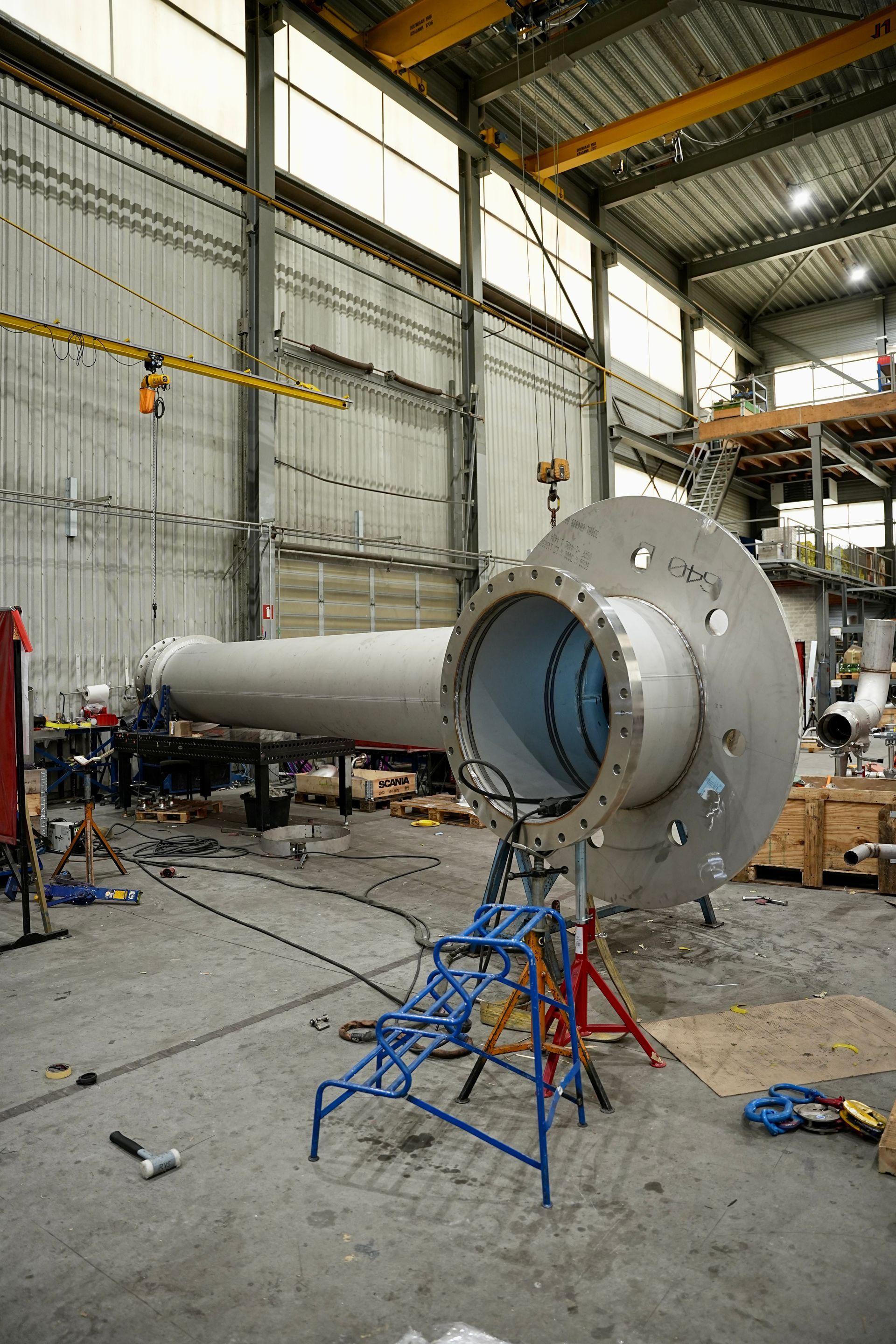 Een grote metalen buis met een ronde flens rust op een blauwe steun in een hoge industriële werkplaats.