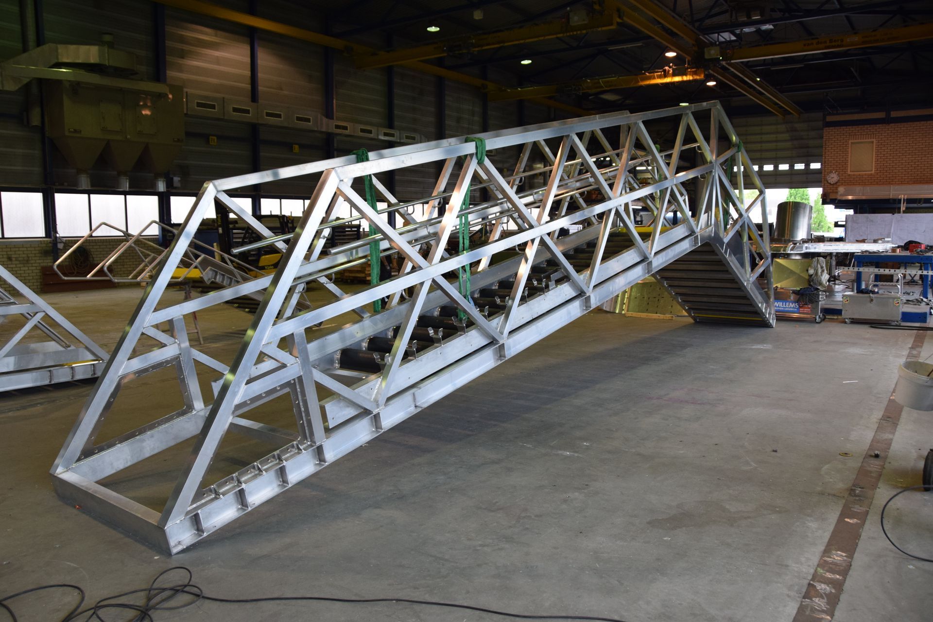Een groot, onafgewerkt metalen brugdeel staat in een felverlicht industrieel magazijn.