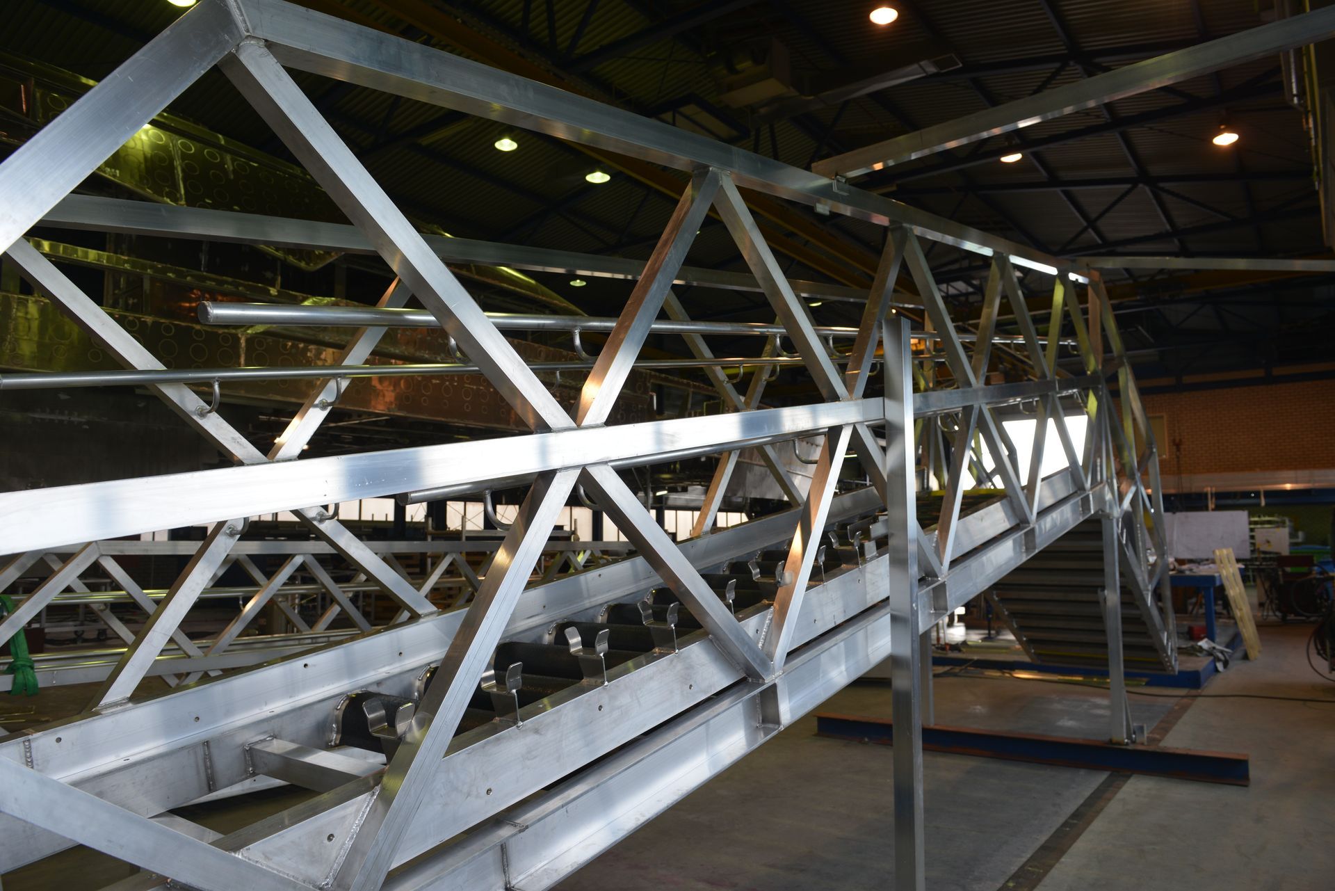 Een zilverkleurig metalen voetgangersbrugframe, opgebouwd uit vakwerksecties, staat in een grote, overdekte industriële werkplaats.