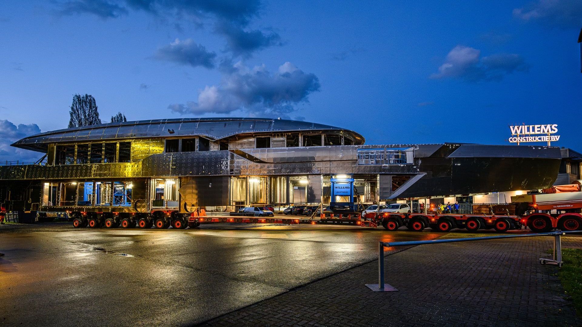 Een groot jachtbouw casco opbouw wordt 's nachts door vrachtwagens verplaatst.