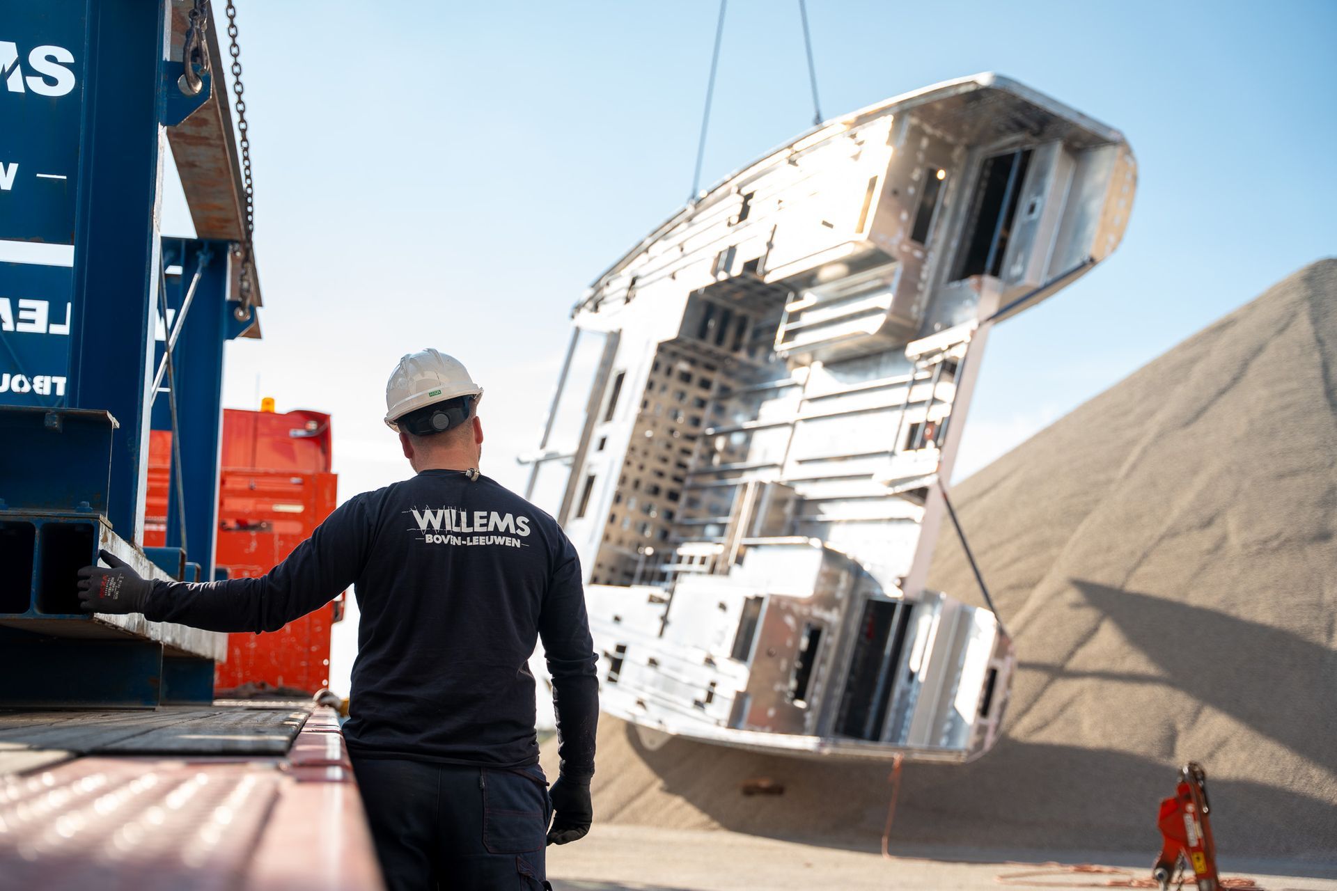 Een arbeider met een veiligheidshelm kijkt toe hoe een groot, metalen industrieel onderdeel door een kraan wordt opgetild nabij een hoop grind.