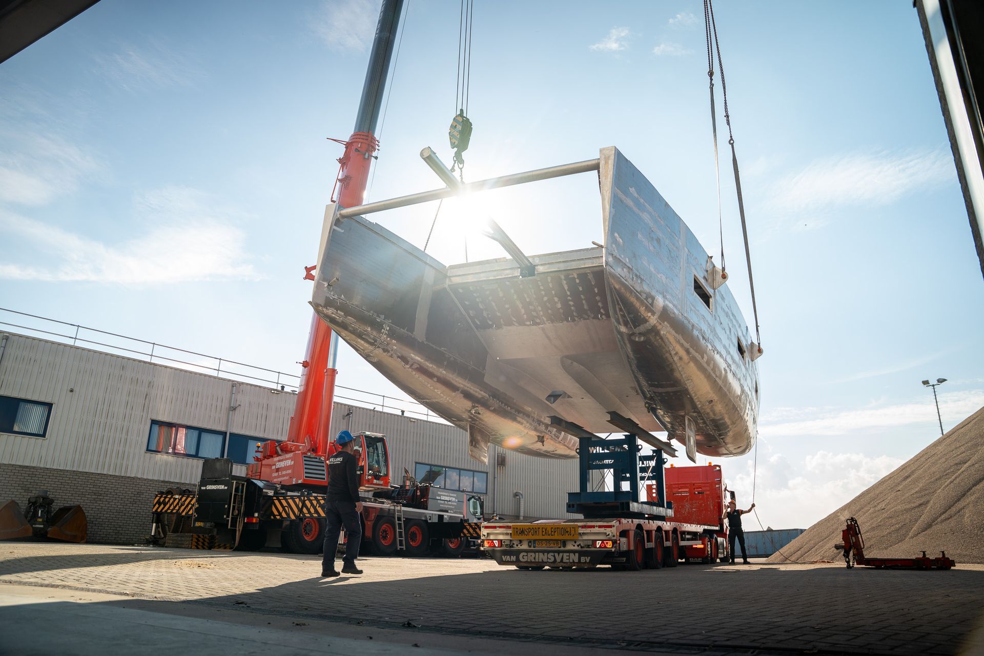 Een kraan tilt een grote metalen bootromp op een transporttrailer op een grindterrein in de felle zon.