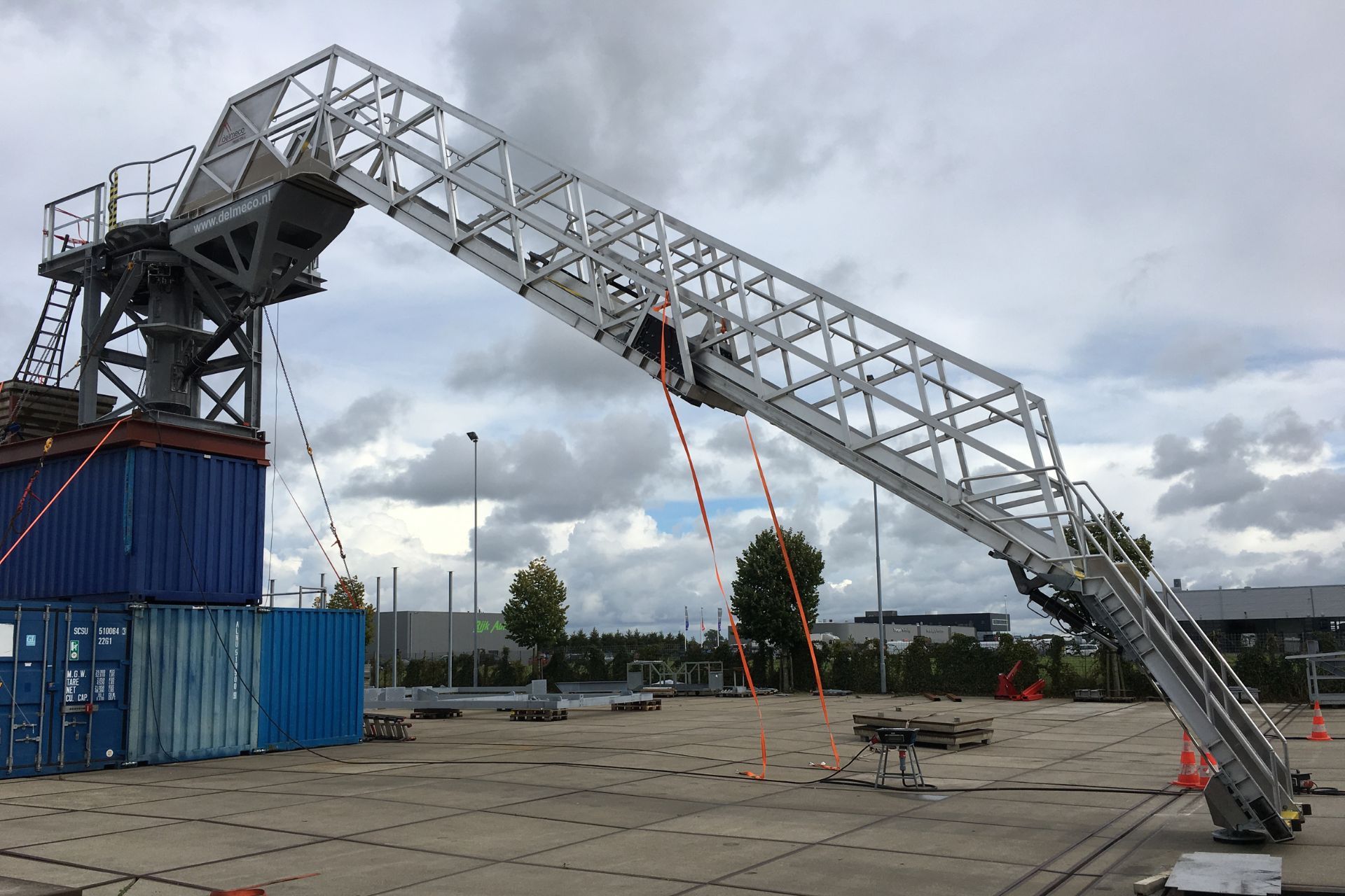 Een grote, metalen, industriële hijsarm, schuin naar beneden gericht, ondersteund door gestapelde blauwe zeecontainers.