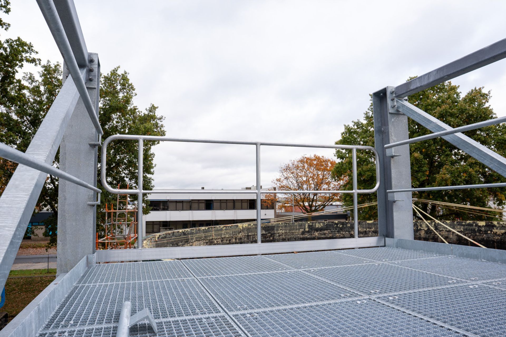 Een laag camerastandpunt van een grijs metalen industrieel platform met een veiligheidsleuning, met uitzicht op bomen en een gebouw in de verte.