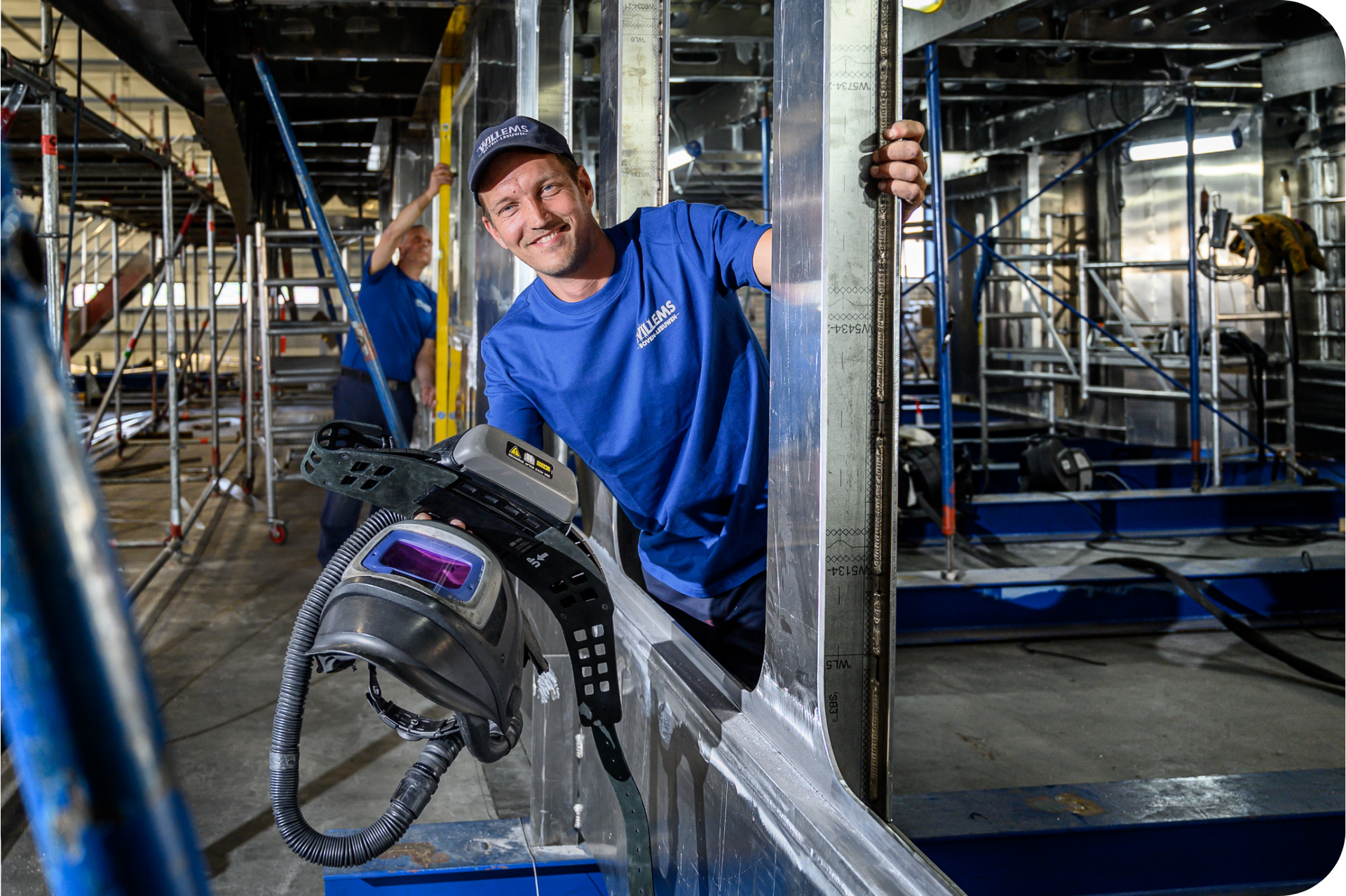 A man in a blue shirt stands in a factory with a welding mask in his hand. By Willems Boven-Leeuwen.