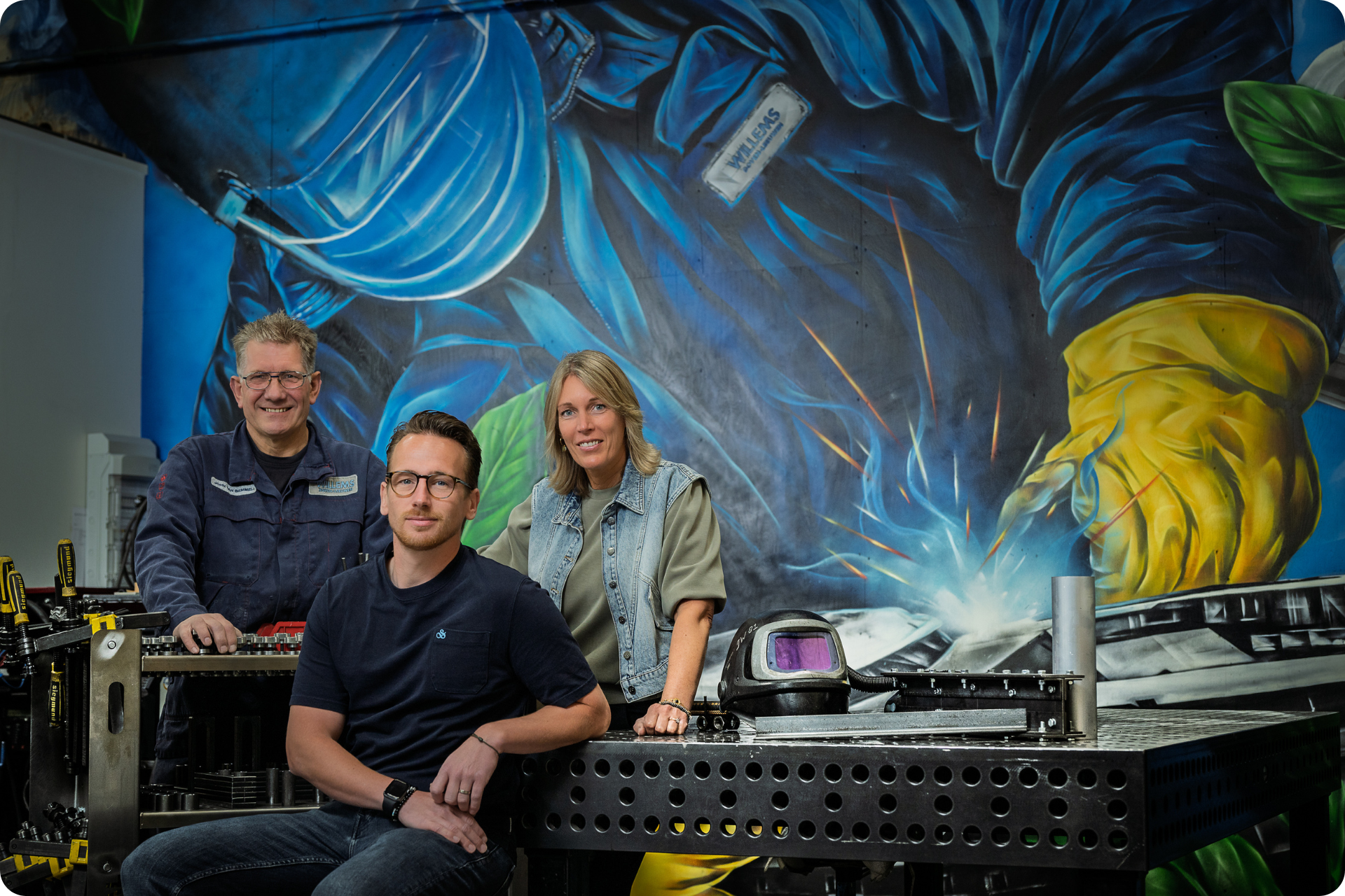 Drie mensen staan ​​voor een grote blauwe muurschildering van een lasser, die zich achter een metalen werkbank in een werkplaats bevindt.