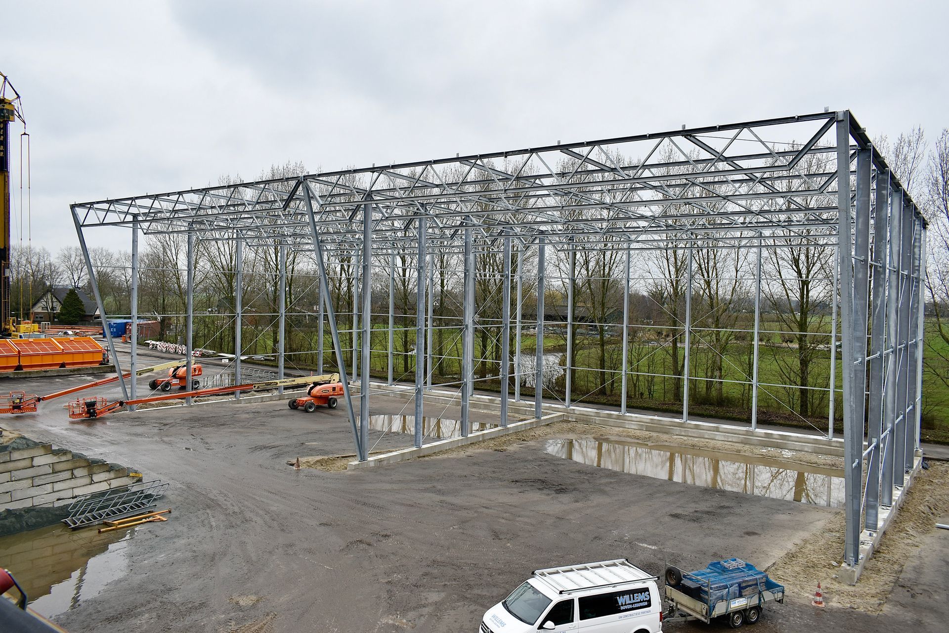 Een wit busje staat geparkeerd voor een groot metalen bouwwerk in aanbouw. Staalconstructie APH Hoogblokland. Willems Boven-Leeuwen.