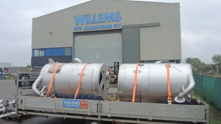 Voor een gebouw staat een grote roestvrijstalen tank op een aanhanger, waarop willems staat