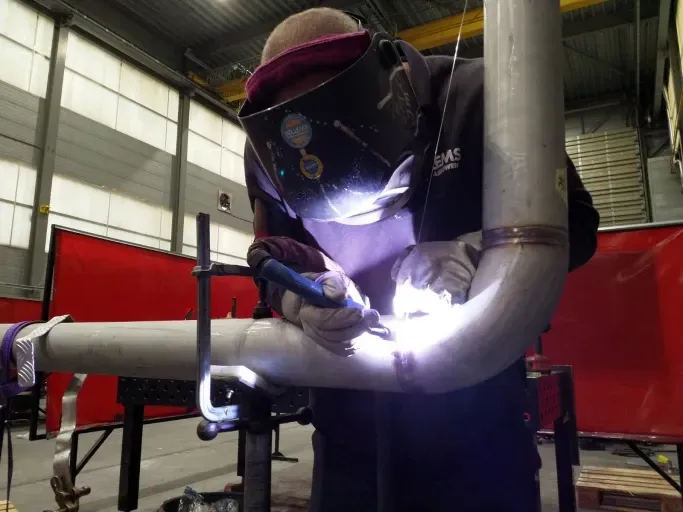 Een man met een lasmasker is een pijp aan het lassen in een fabriek. Willems Piping & Equipment