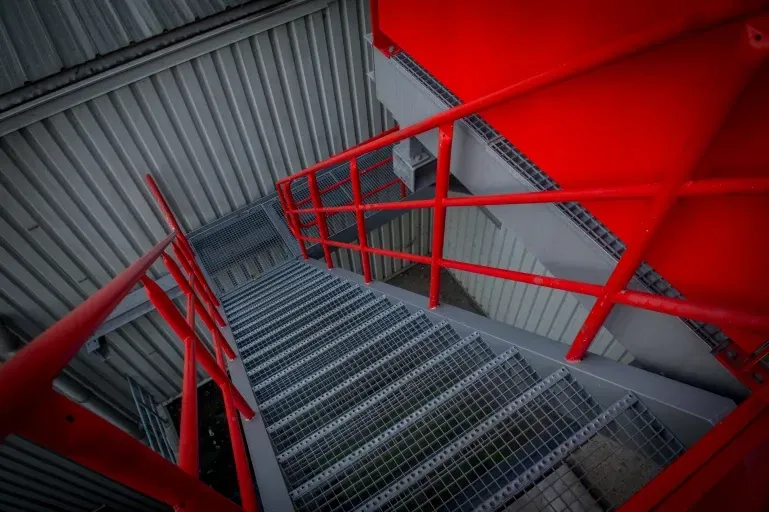 Een metalen trap met een rode reling in een gebouw. Door Willems Boven-Leeuwen.