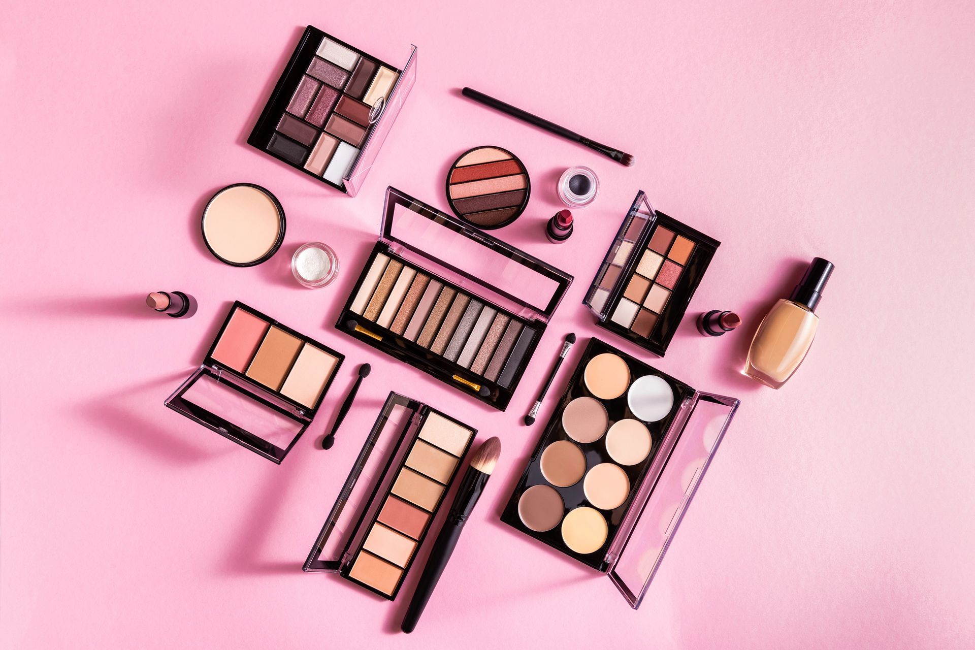 Productos de maquillaje, incluyendo paletas, bases y labiales, dispuestos sobre una superficie rosa.