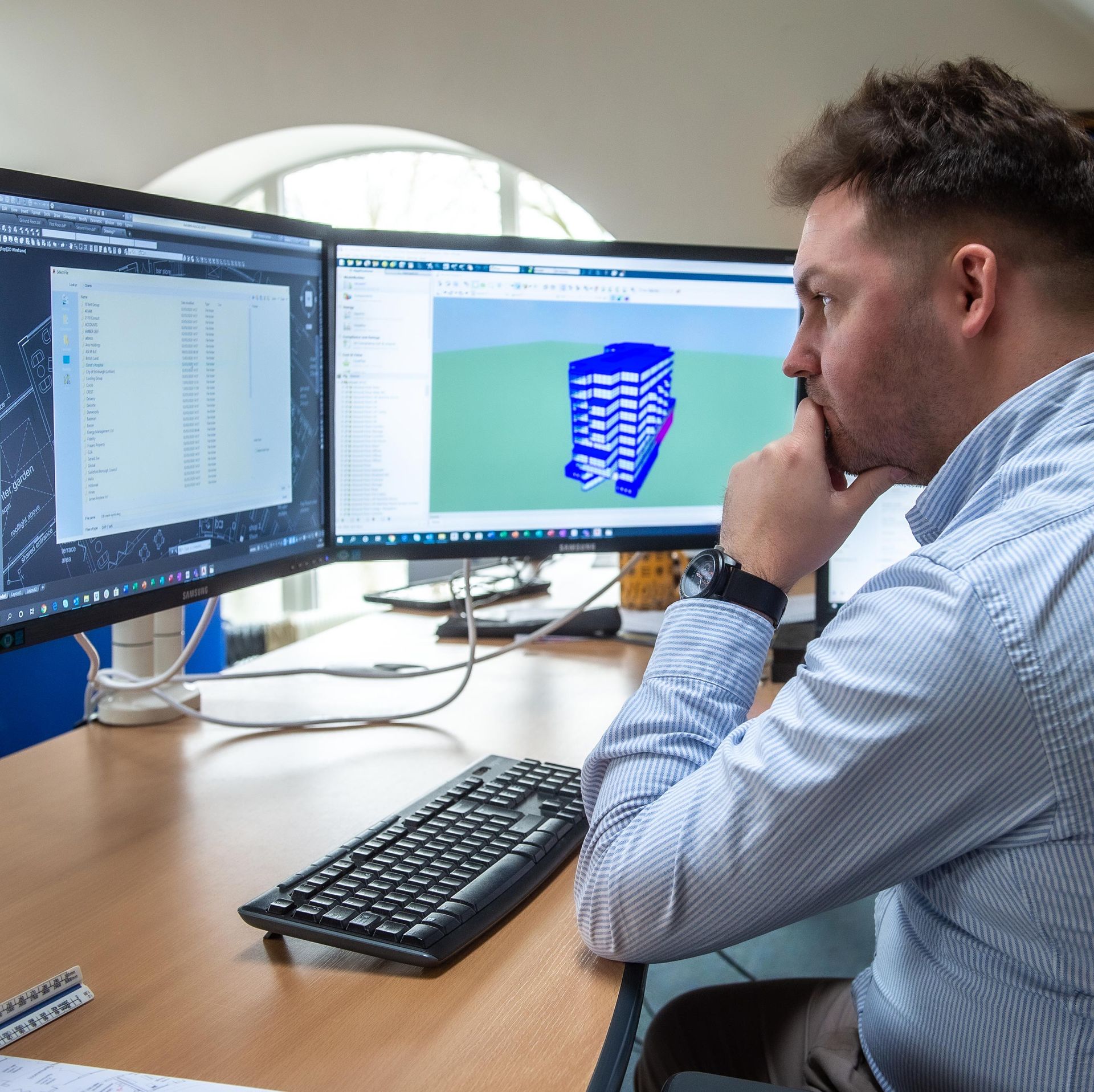 Man working at a desk with two computer monitors displaying architectural designs.