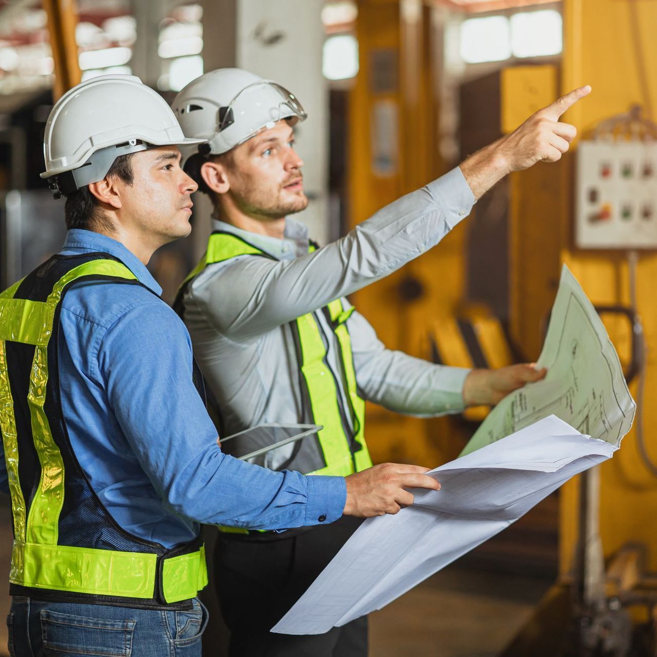 Two construction workers in hard hats and vests examining blueprints at a worksite. One points.
