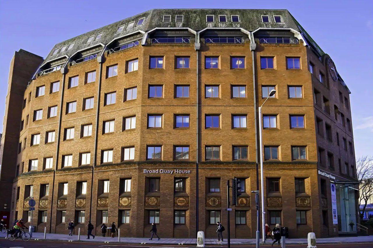 A large brick office building with some figures walking along the pavement in front of it. 