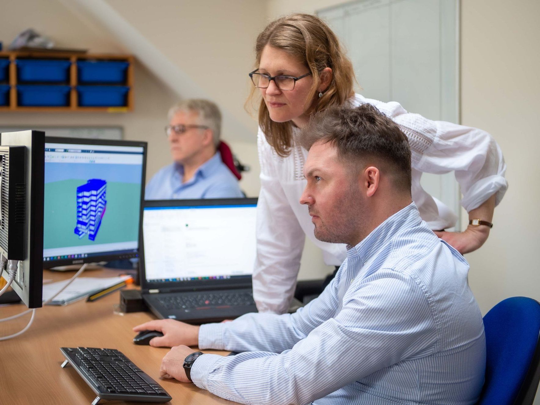 Two people looking at a computer screen displaying a 3D model; another person in background.