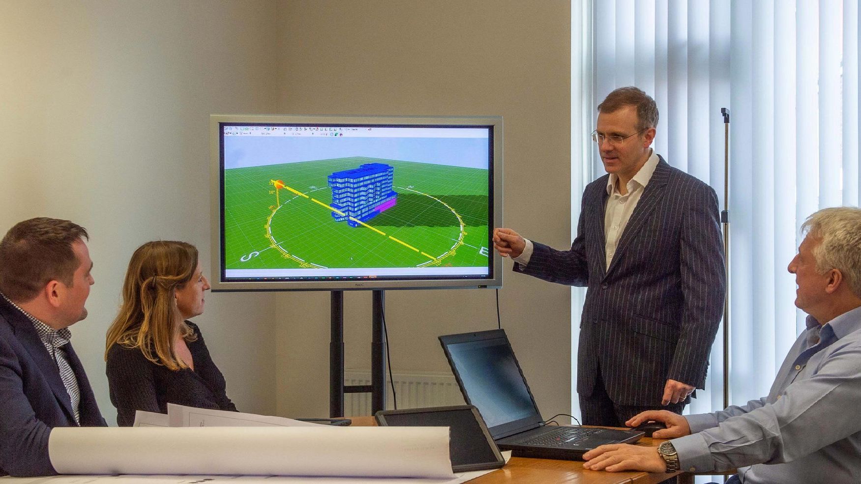 Group in office; man points to screen with 3D model; two others watch, one uses a laptop, plans on the table.