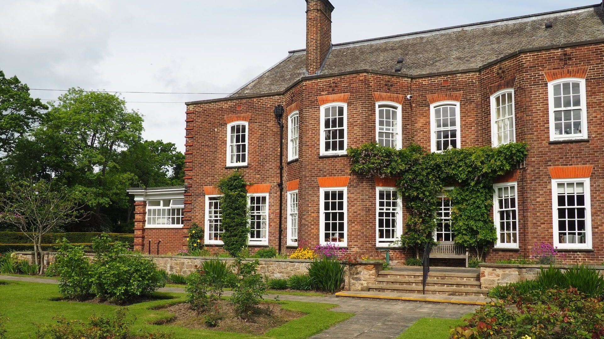 Two-story brick building with white-framed windows, surrounded by a garden and green foliage.