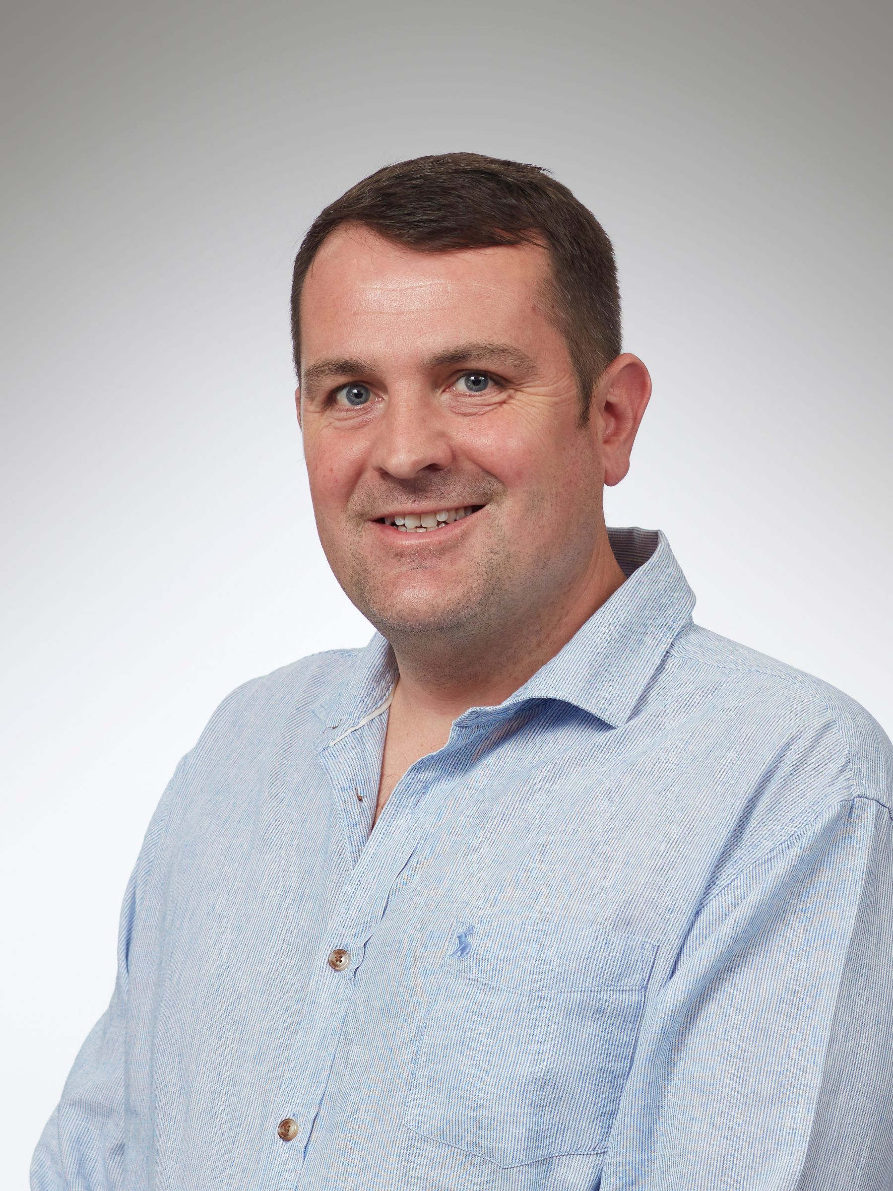 Man with short brown hair smiles, outdoors, wearing a light blue collared shirt.