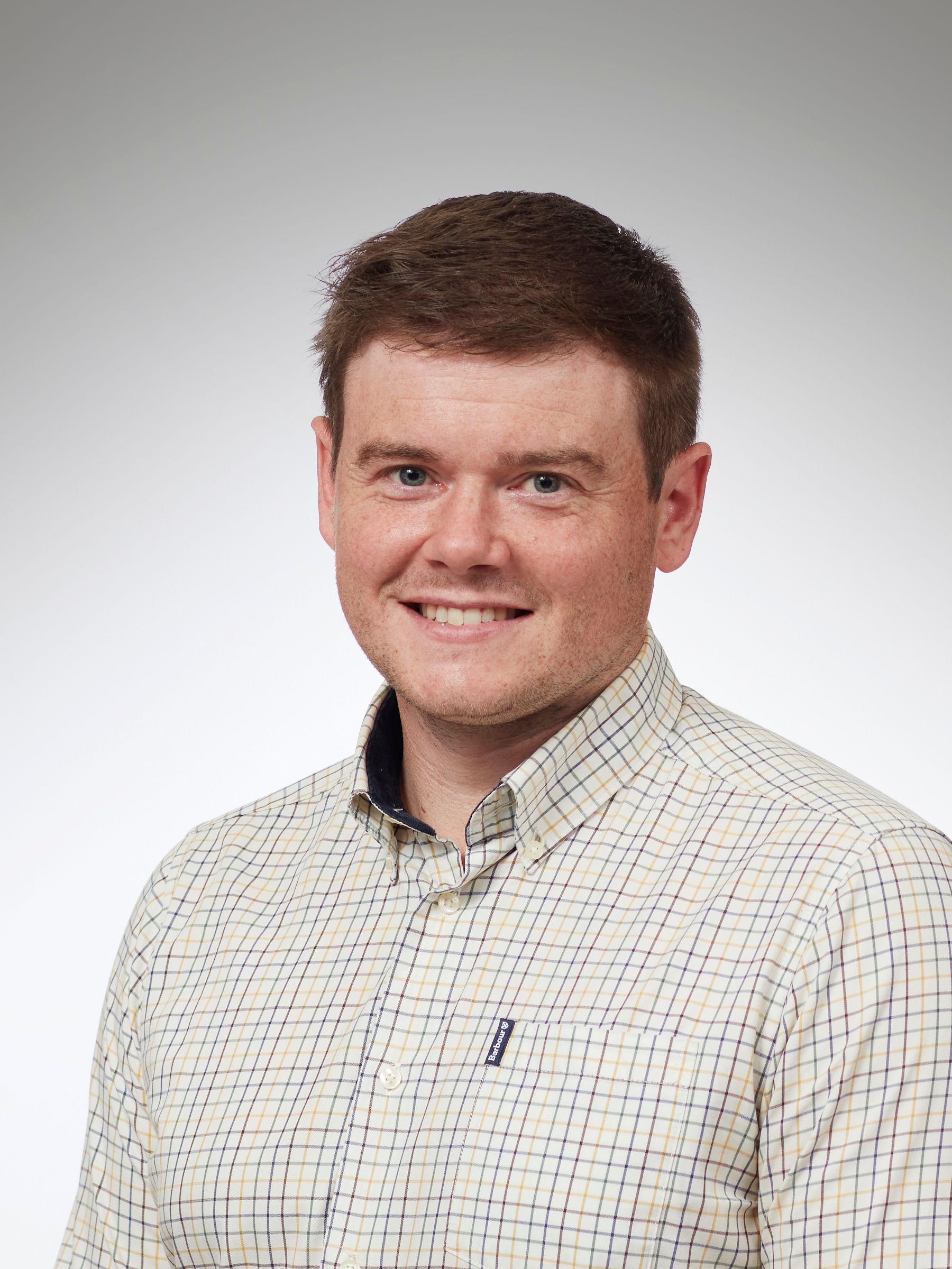 Man in purple button-down shirt smiles outside. Green trees and water are in the background.
