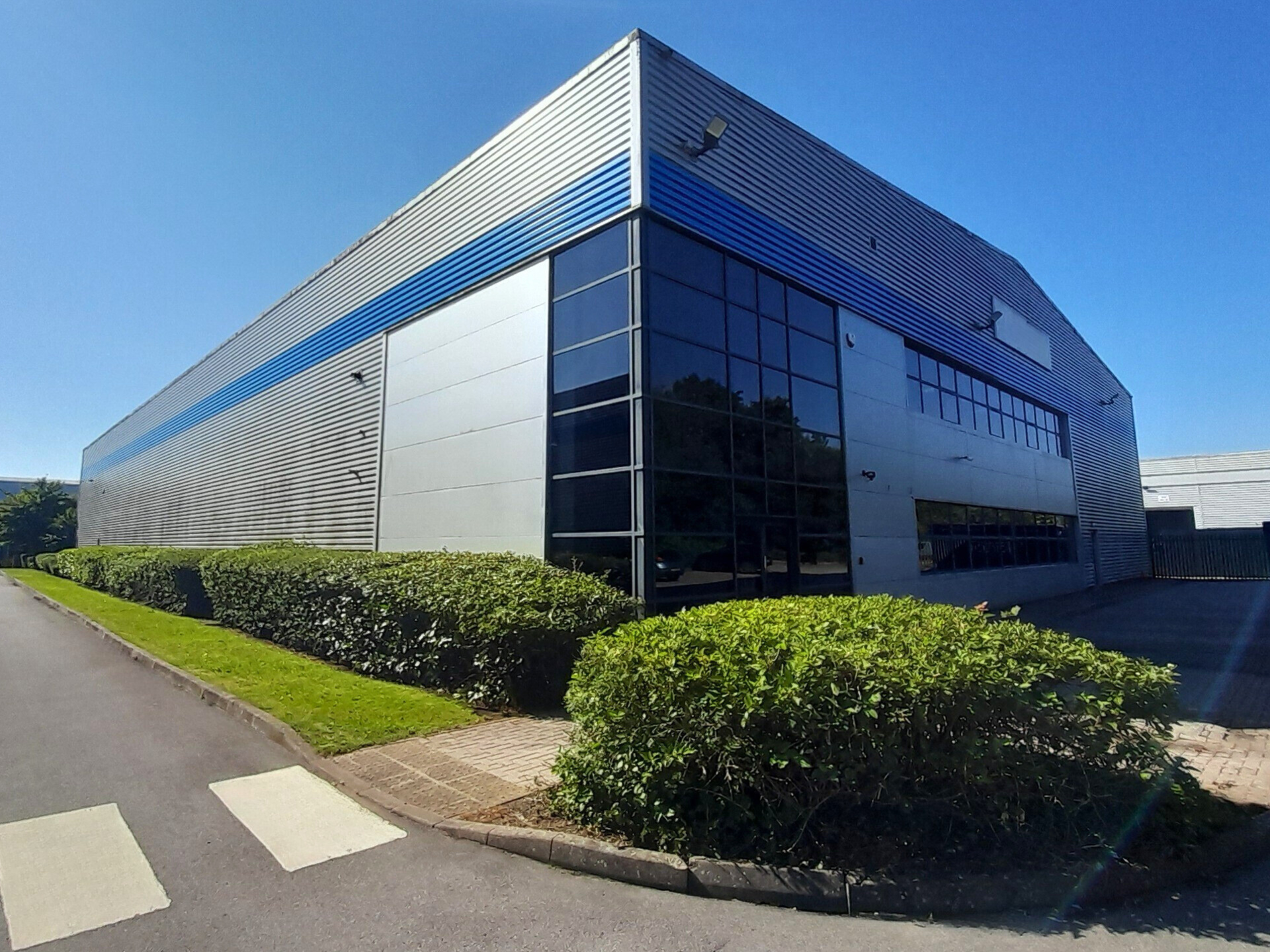 Modern industrial building with gray and blue facade, large windows, and manicured bushes under a blue sky.