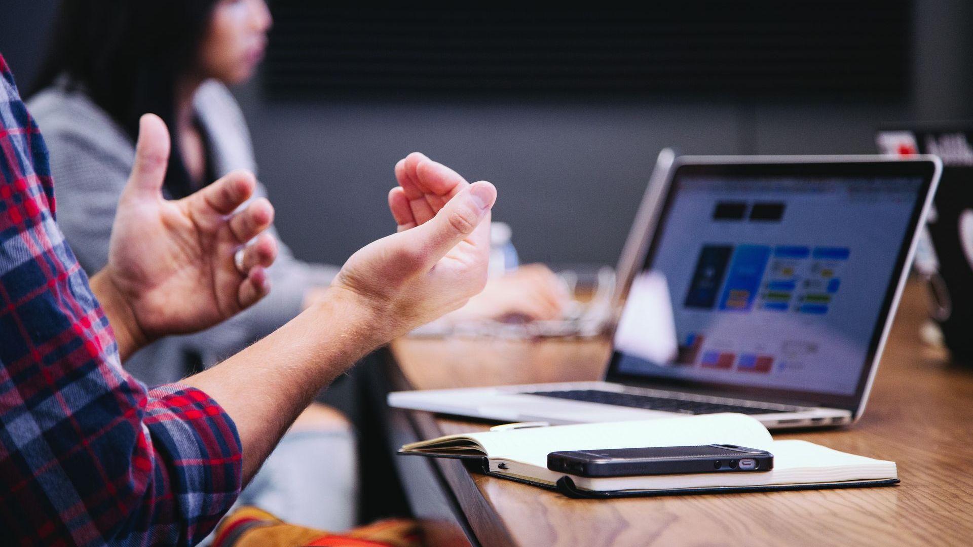 Person gesturing with hands during a meeting, laptop and notebook on table.