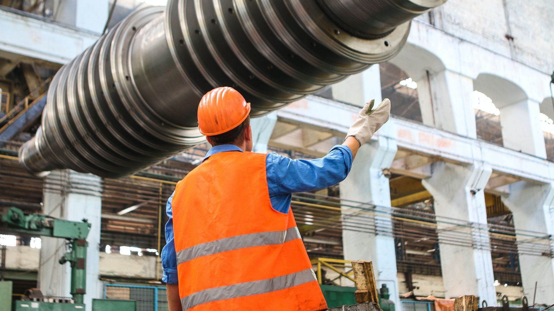 Worker in orange vest and hard hat inspects large metal machinery in a factory setting.