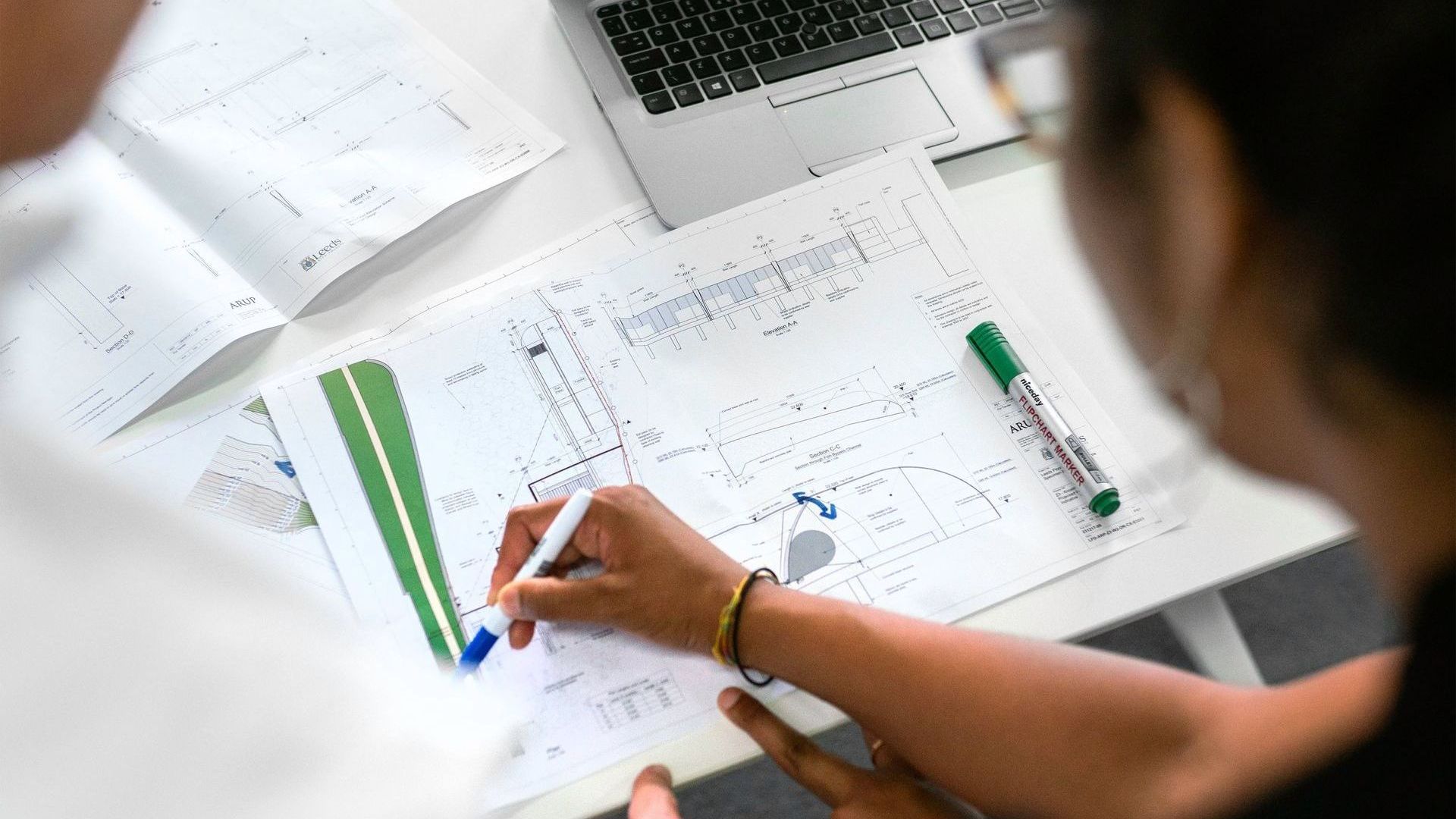 Two people working on blueprints, writing with a pen. Laptop and open book are on the table.
