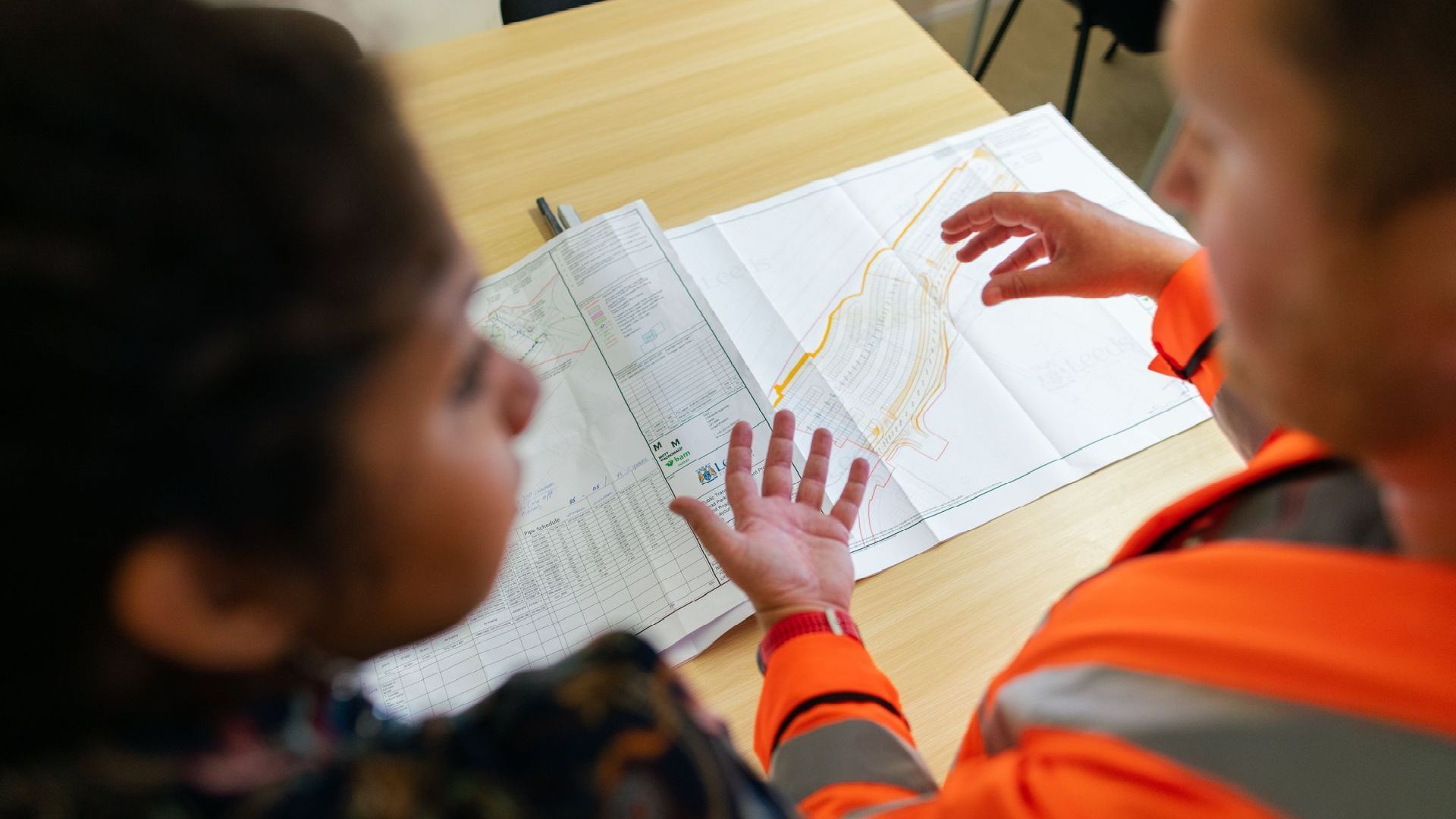 Two people reviewing a detailed map on a table; one points at the map with orange safety gear.