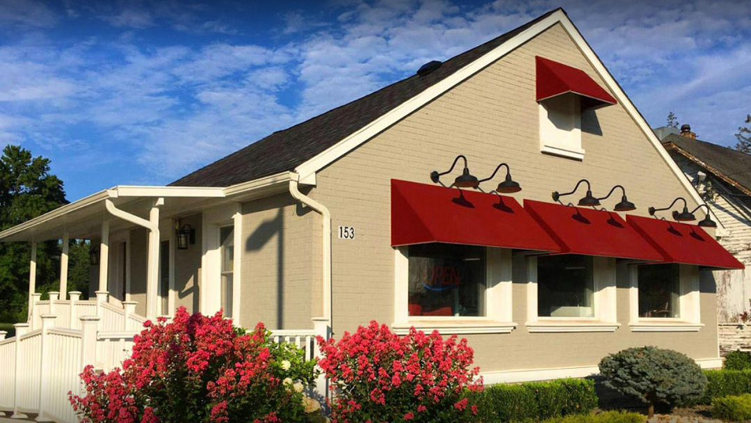 Tan building with red awnings and outdoor lighting, white porch and fence.