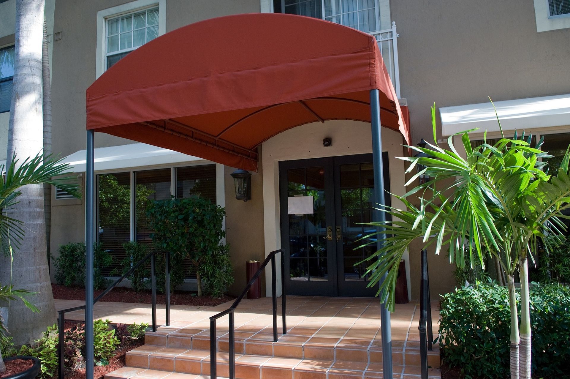 Exterior of a building with red awning over the entrance, steps, bushes, and palm trees.
