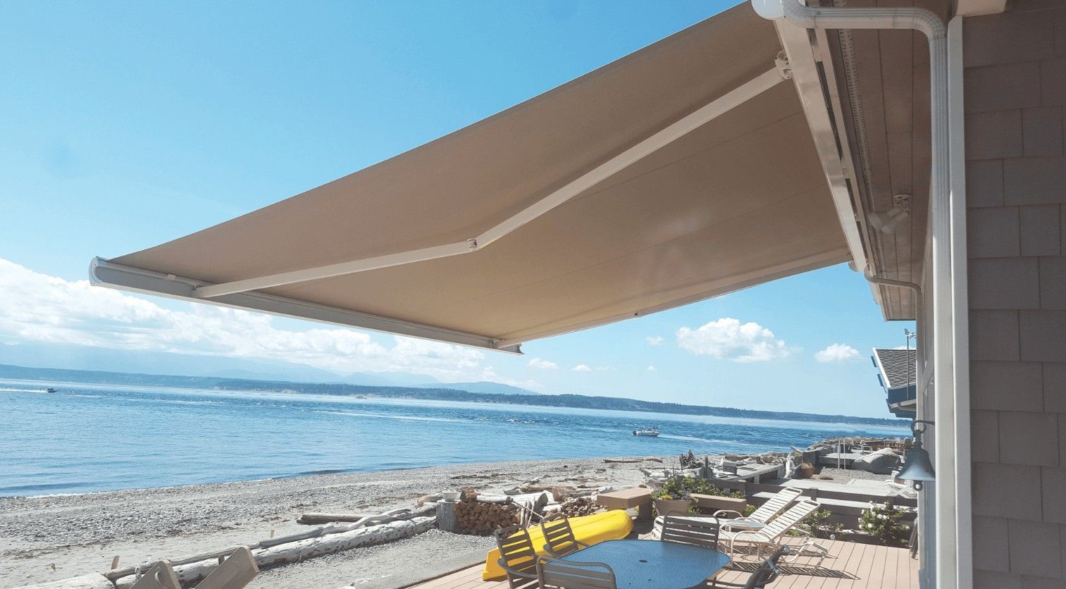 Beige retractable awning extends from a white house overlooking a beach and ocean. Sunny day.