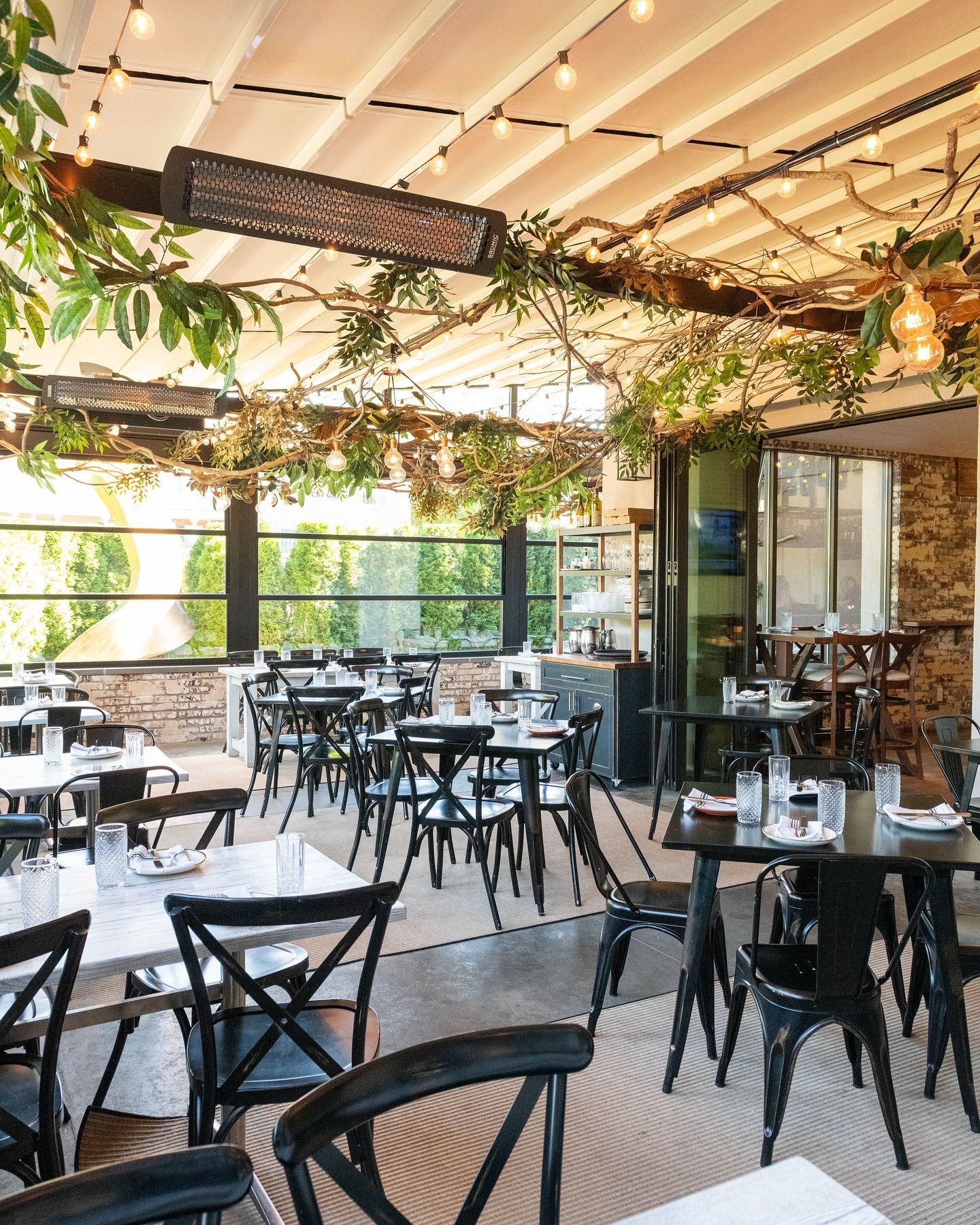 Outdoor restaurant patio with tables, chairs, and leafy vines. Overhead heaters and string lights visible.