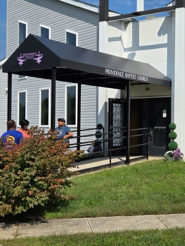 Providence Baptist Church entrance with awning, people gathered outside. Ramp, black door, green lawn.