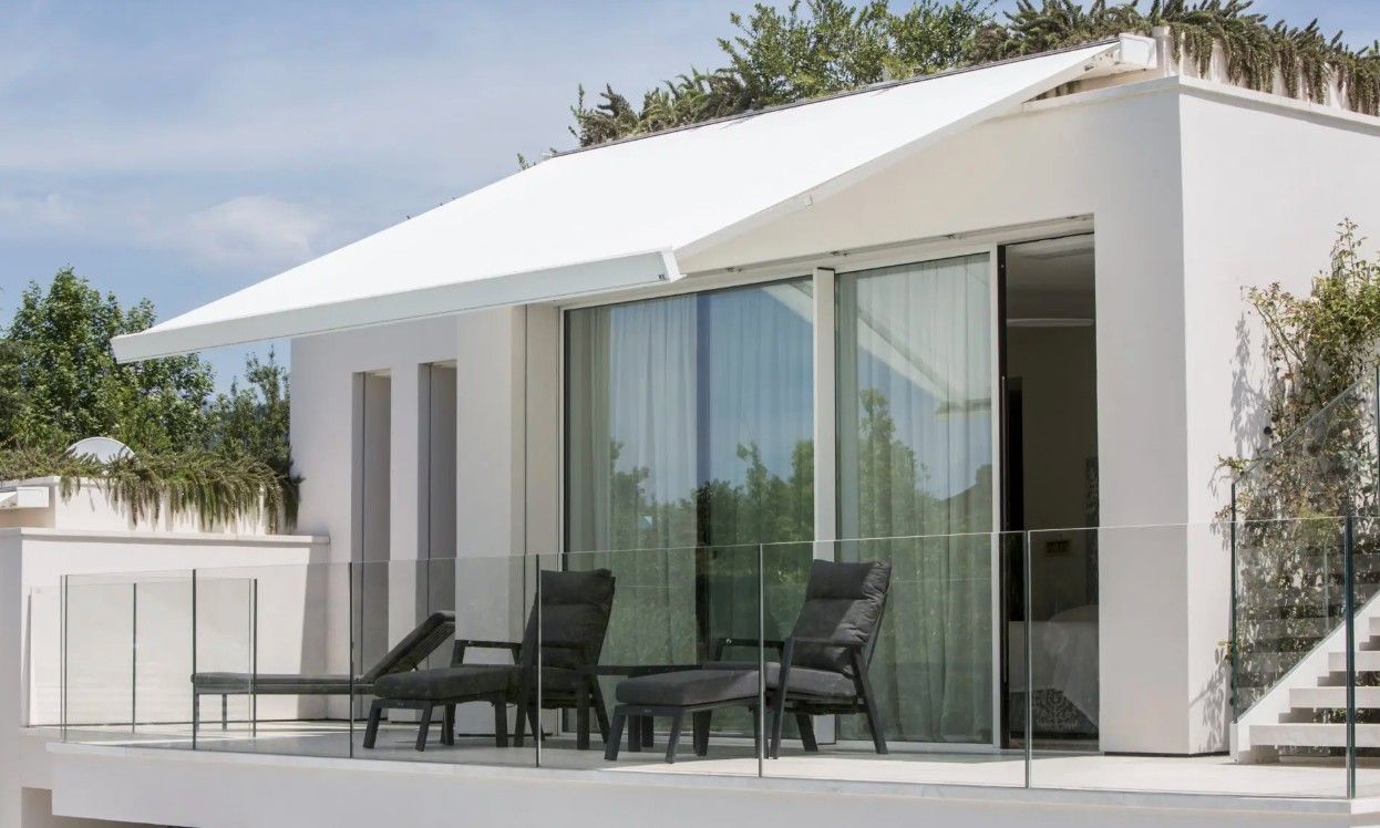 White building with large glass doors, outdoor seating, awning, and glass railing.