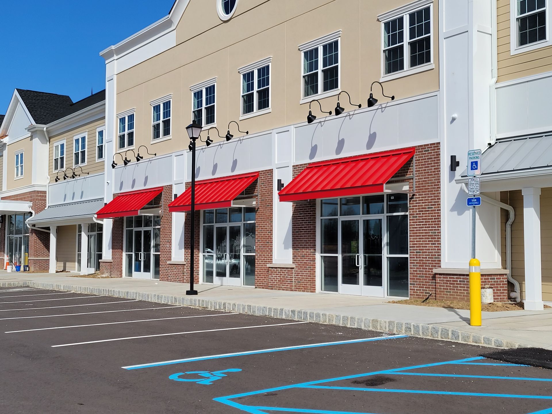 Exterior view of a shopping center with red awnings, glass storefronts, and parking spaces.