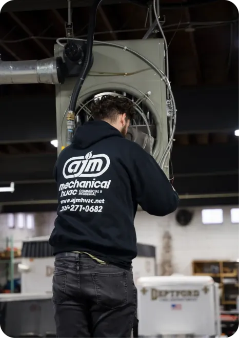 A man in a hoodie (AJM Mechanical) is working on an industrial HVAC unit. He's in a warehouse.