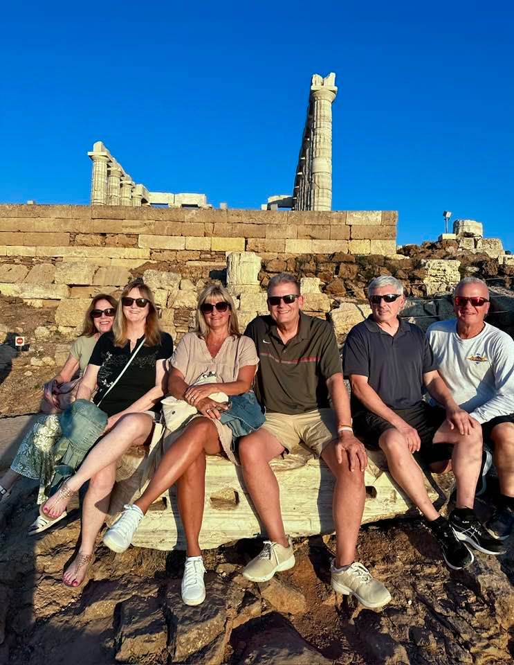 Group of six people sit on ancient stone ruins in front of a blue sky and tall pillar.