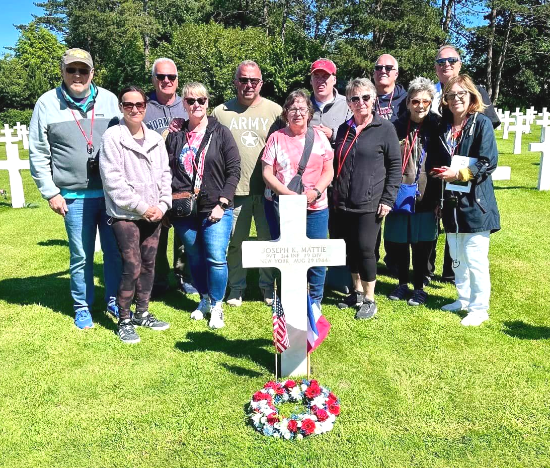 Group stands at a grave in a white-cross cemetery. Red, white, and blue decorations. Green grass and trees in background.
