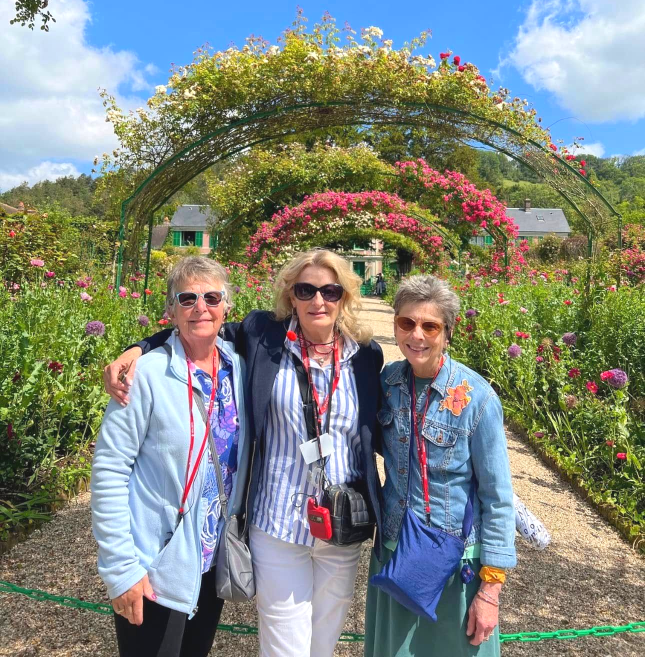 Three people pose under rose archway; garden path, sunny day.