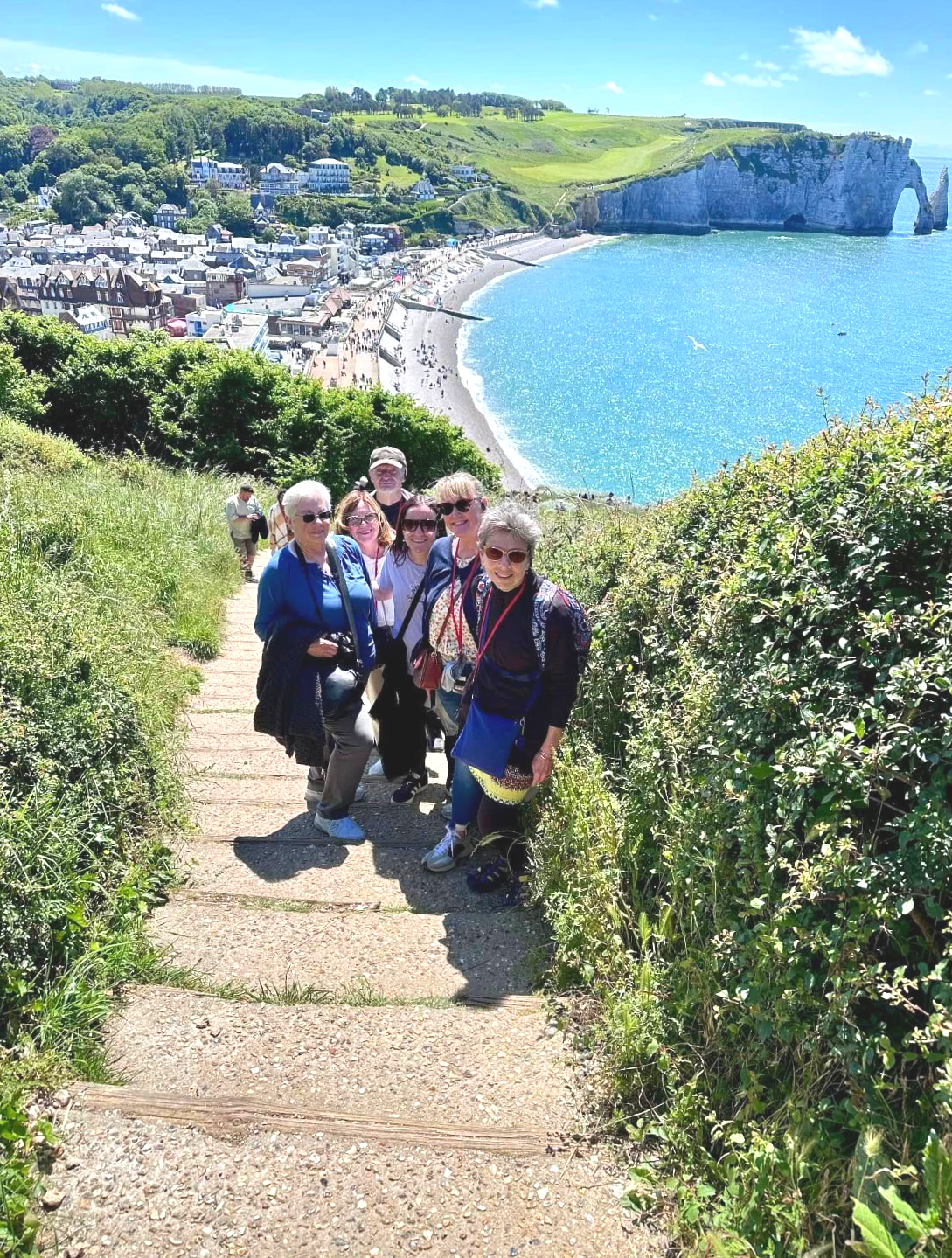 Group of people on a path overlooking a coastal town and cliffs; blue water, sunny day.