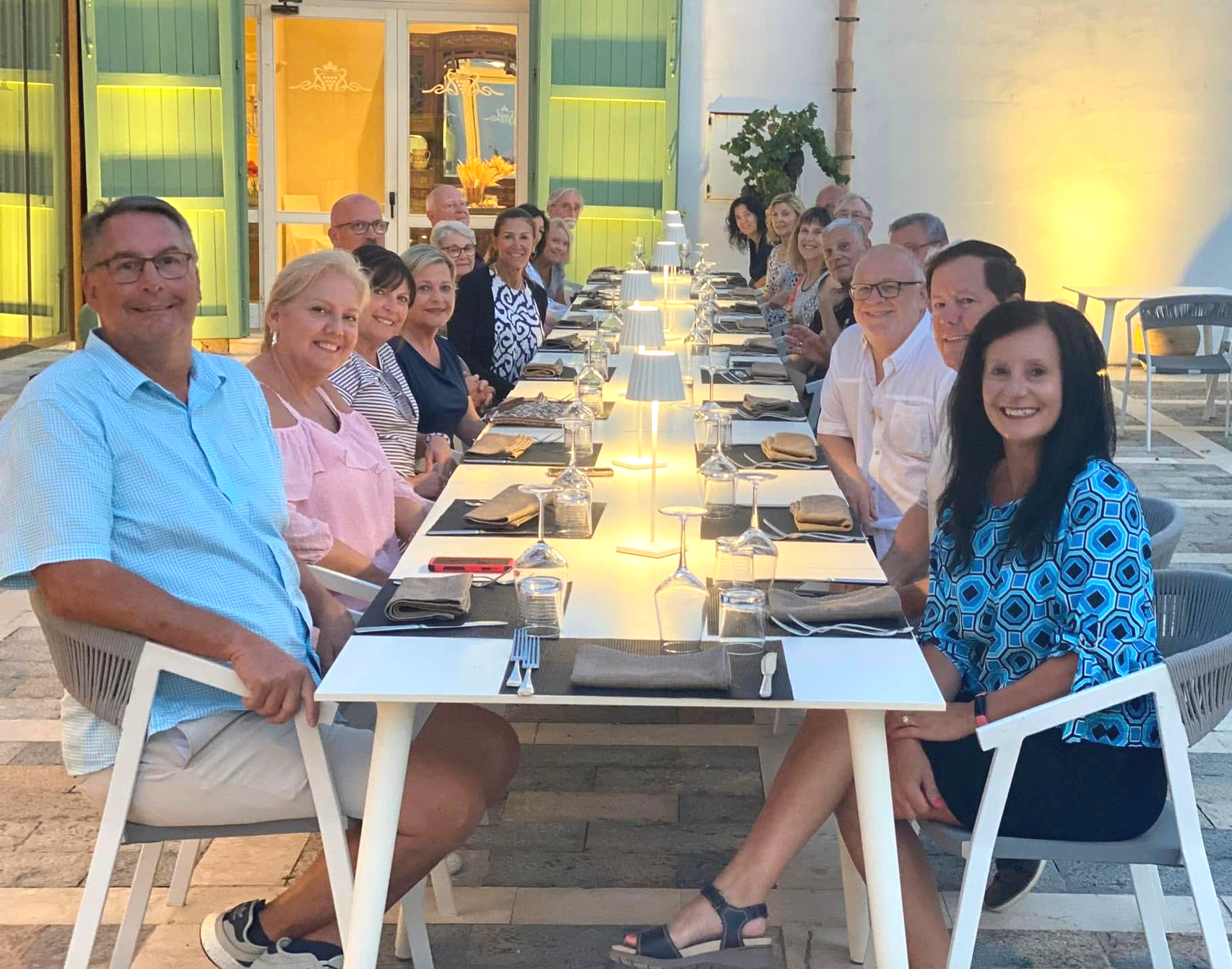 Group of people seated at a long outdoor dining table; some smiling. Bright lighting, neutral colors, modern setting.